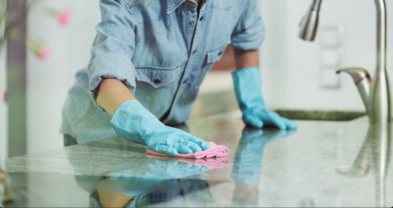 The image appears to show a person cleaning a kitchen countertop. They are wearing blue rubber gloves and are using a pink cloth to wipe down the glossy stone surface. The kitchen has a modern feel with bright lighting, and there are pink flowers in the background, suggesting a well-kept, hygienic home environment.